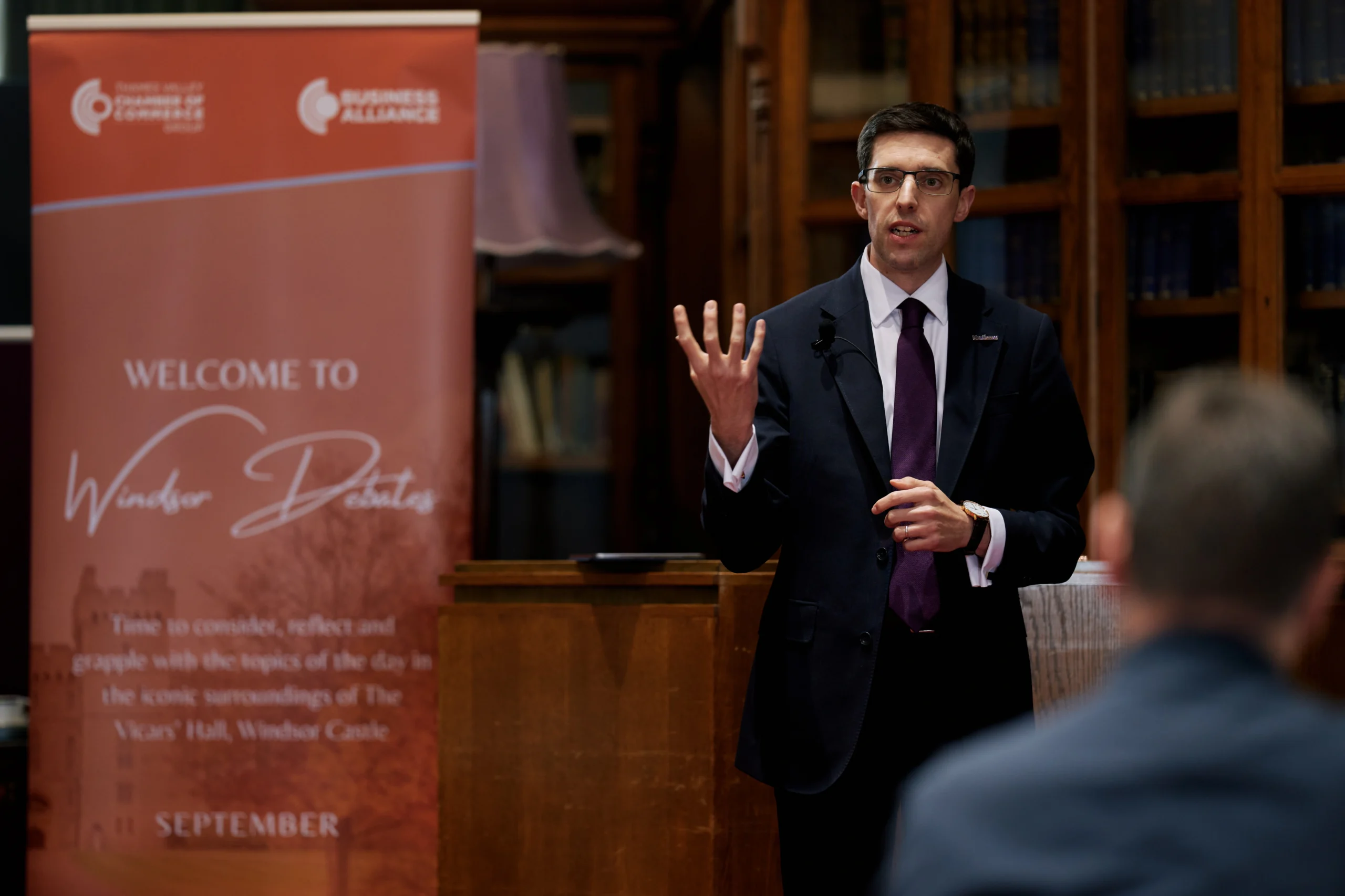 Man in a dark suit and glasses speaks at a podium, gesturing with his left hand in a library-like room with a red banner beside him.