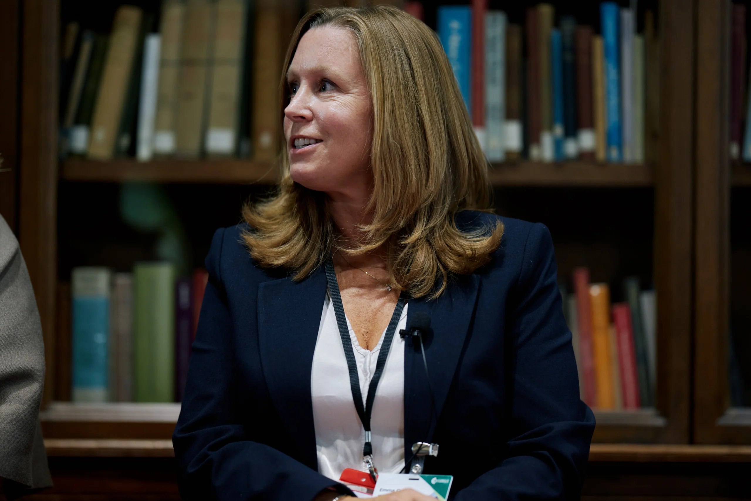 Professional woman in a navy blazer sits in a library, speaking with a lapel microphone clipped to her jacket.