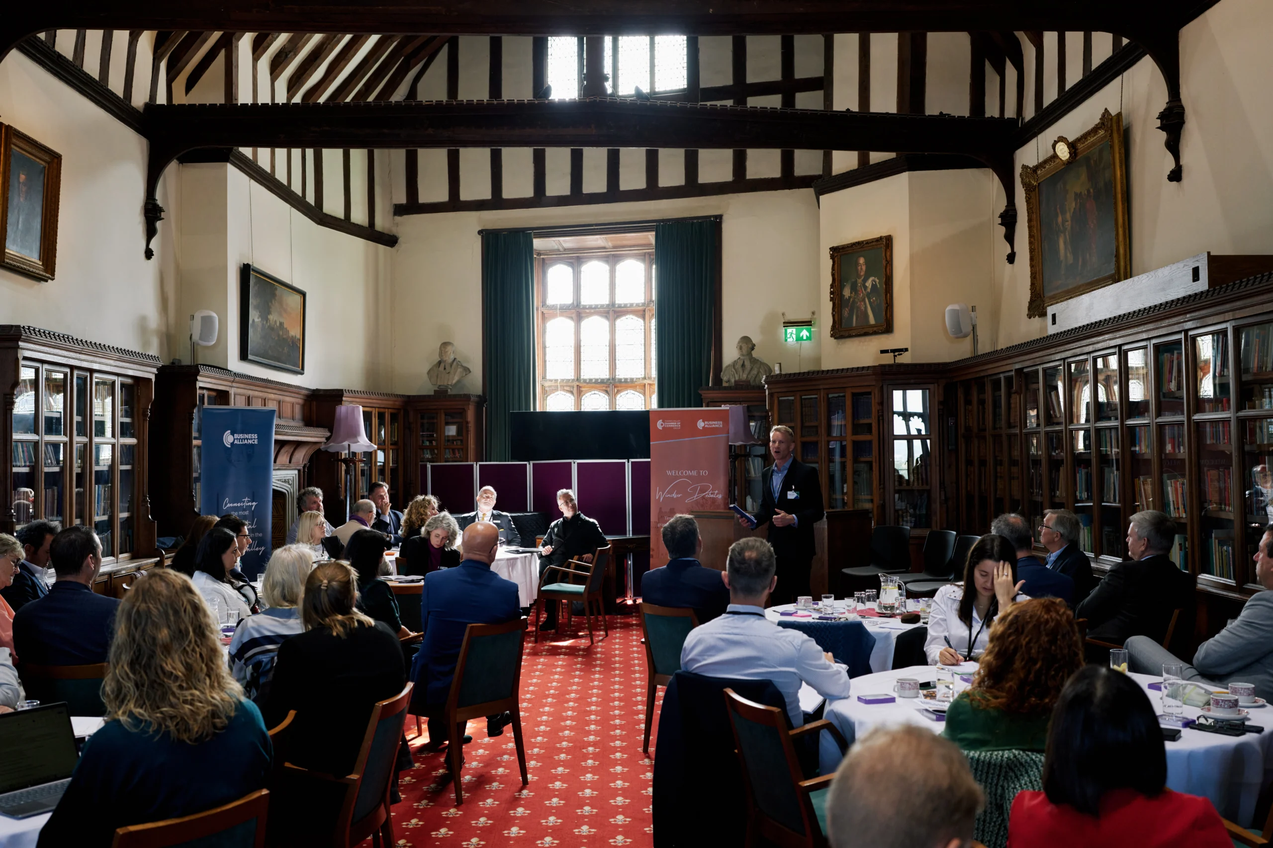Conference in an ornate library: attendees seated around round tables while a speaker stands at a lectern at the front, banners visible behind him.