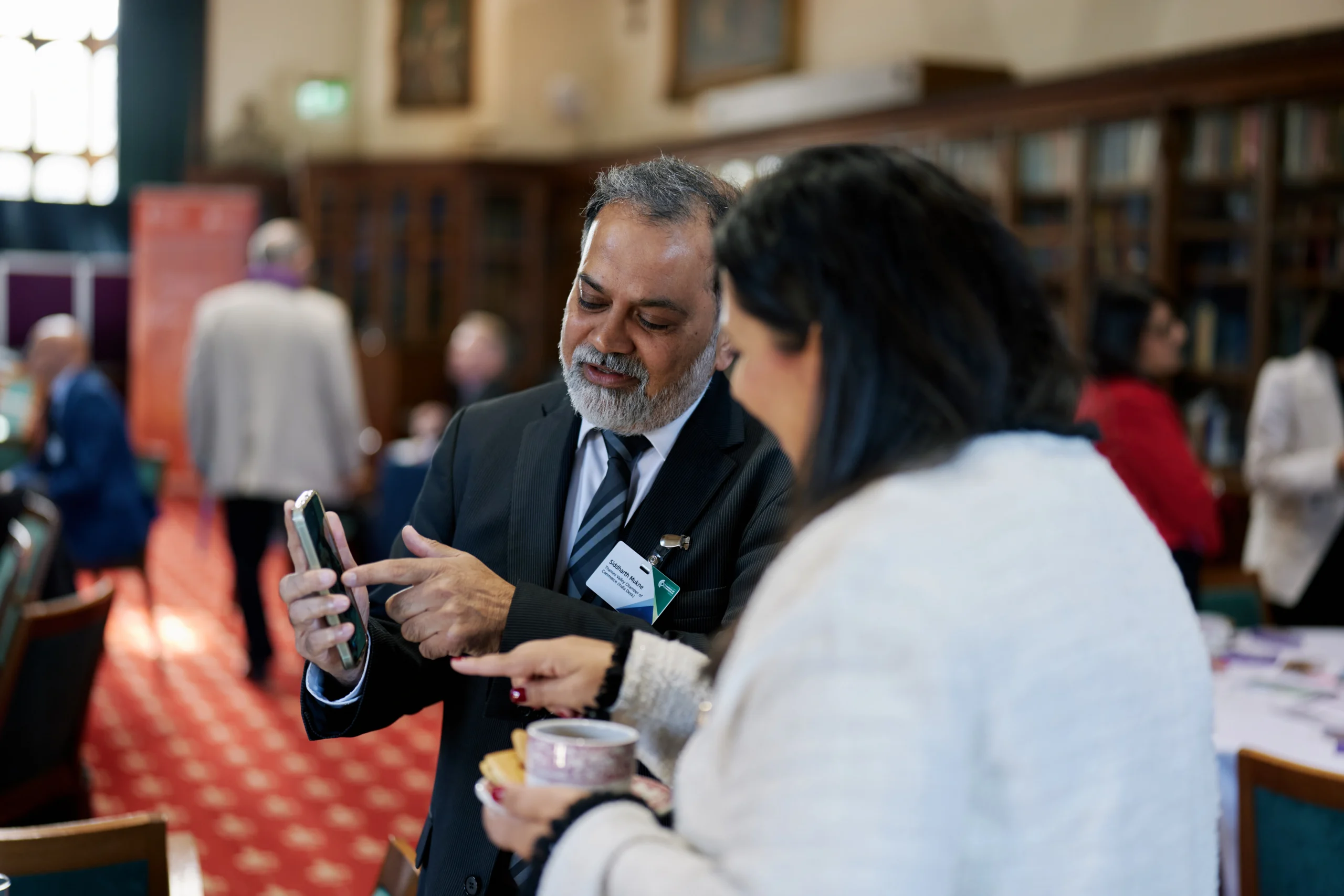 Man in a dark suit showing his phone to a woman at a social event, with name badge visible on his chest and a library-style backdrop.