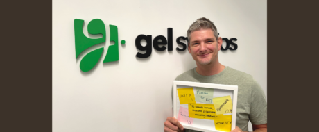 Smiling man in a gray shirt holds a white frame filled with yellow sticky notes in front of a wall with a green company logo and text behind him.