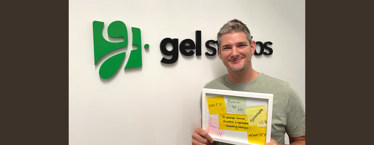 Smiling man in a gray shirt holds a white frame filled with yellow sticky notes in front of a wall with a green company logo and text behind him.