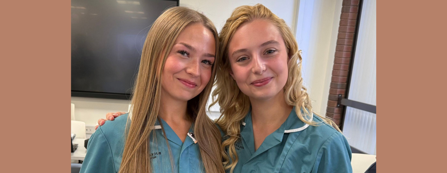 NEW - News item size 27 | Thames Valley Chamber of Commerce Two young women in teal scrubs smiling at the camera in a classroom setting of a training room or lab with a brick pillar nearby.