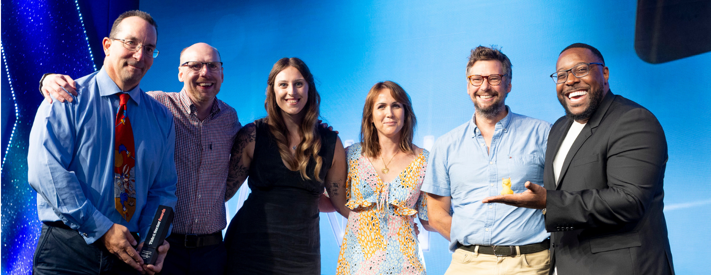 Six adults stand arm-in-arm on a blue-lit stage, smiling for a group photo at a work event or conference point.