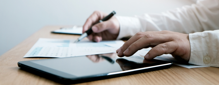 Hands write with a pen on papers while a tablet lies on a wooden desk, suggesting document review or note-taking in progress.