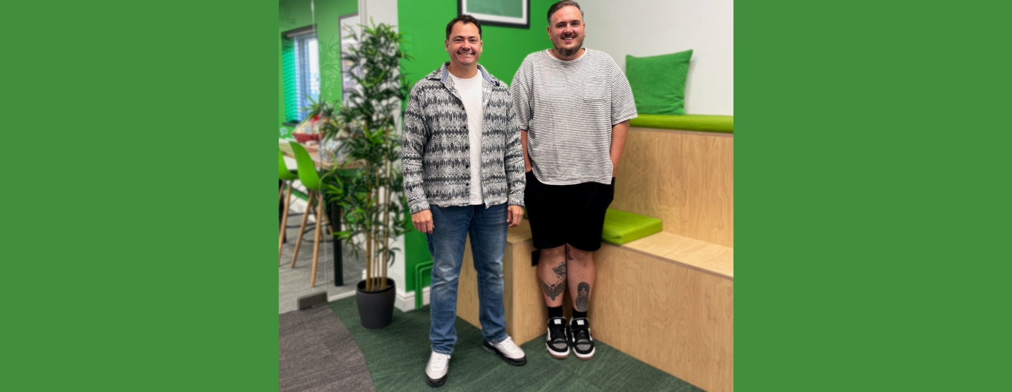 NEW - News item size 77 | Thames Valley Chamber of Commerce Two men posing for a photo indoors near a green-walled area with plants and wooden seating, both smiling at the camera.