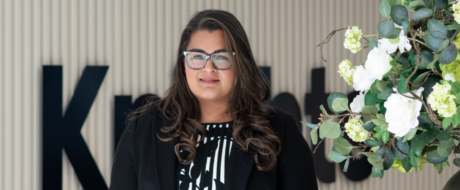 Portrait of a woman with glasses in a black blazer, standing beside a floral arrangement.