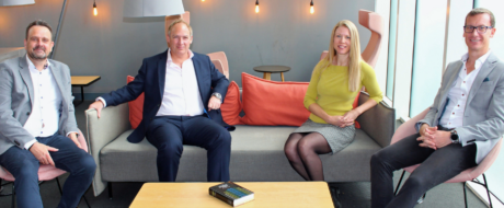 Four business professionals seated on a modern lounge sofa in a bright office, smiling at the camera.