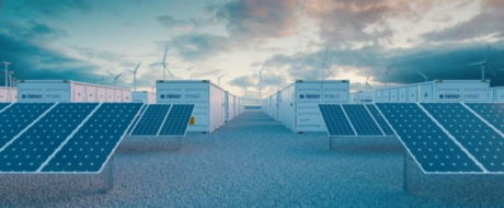 Rows of blue solar panels on concrete risers in a storage facility, with wind turbines visible in the cloudy sky behind them.
