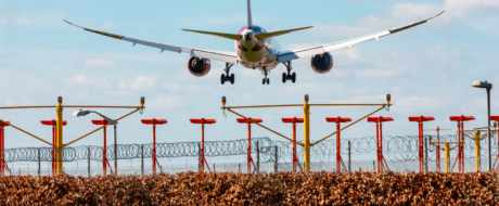 Passenger jet descending toward a runway with landing gear down, seen over a barbed-wire perimeter and red-yellow approach lights.
