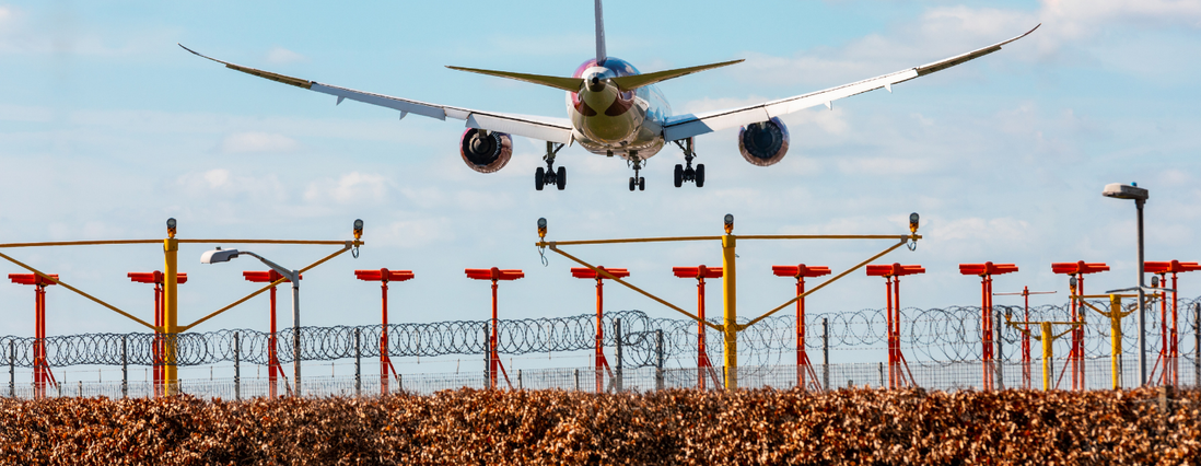 NEW - News item size 85 | Thames Valley Chamber of Commerce Passenger jet descending toward a runway with landing gear down, seen over a barbed-wire perimeter and red-yellow approach lights.