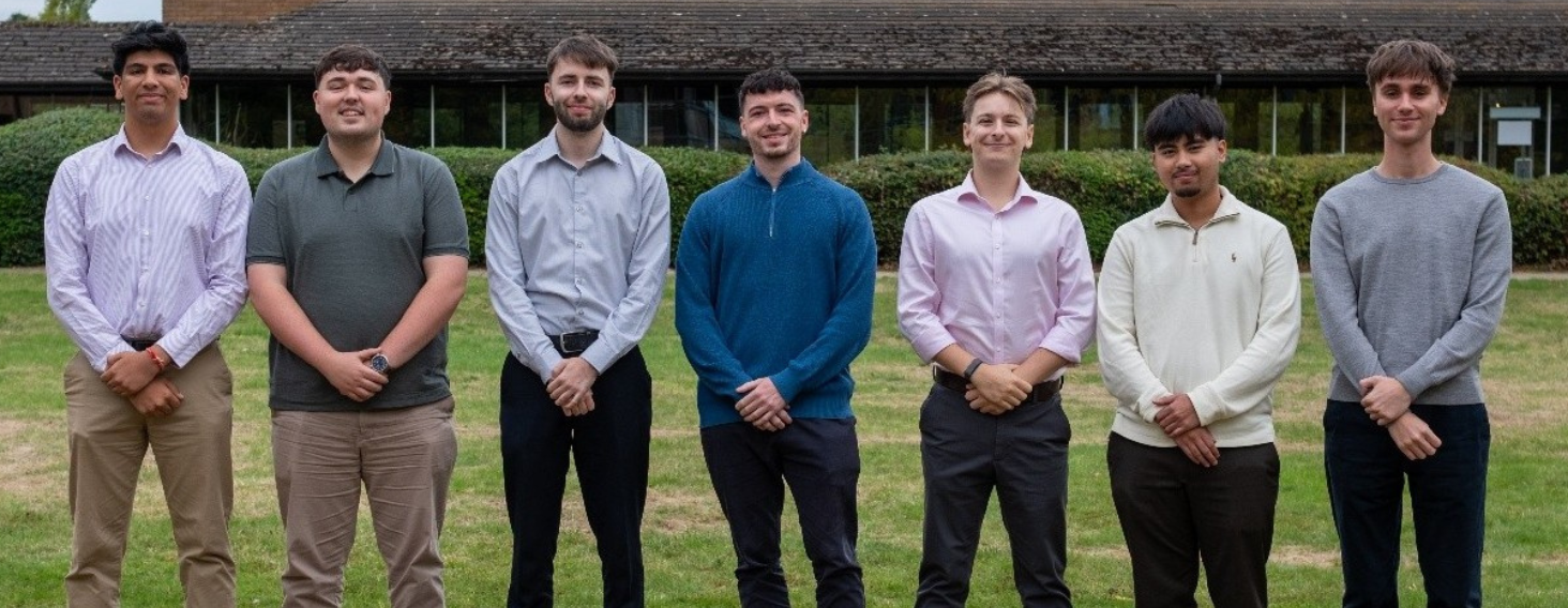 Seven men standing in a row on a grassy field, posing for a group photo on a sunny day.