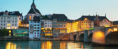 Colorful historic buildings along a calm river at dusk, with an illuminated stone-arch bridge reflecting in the water.
