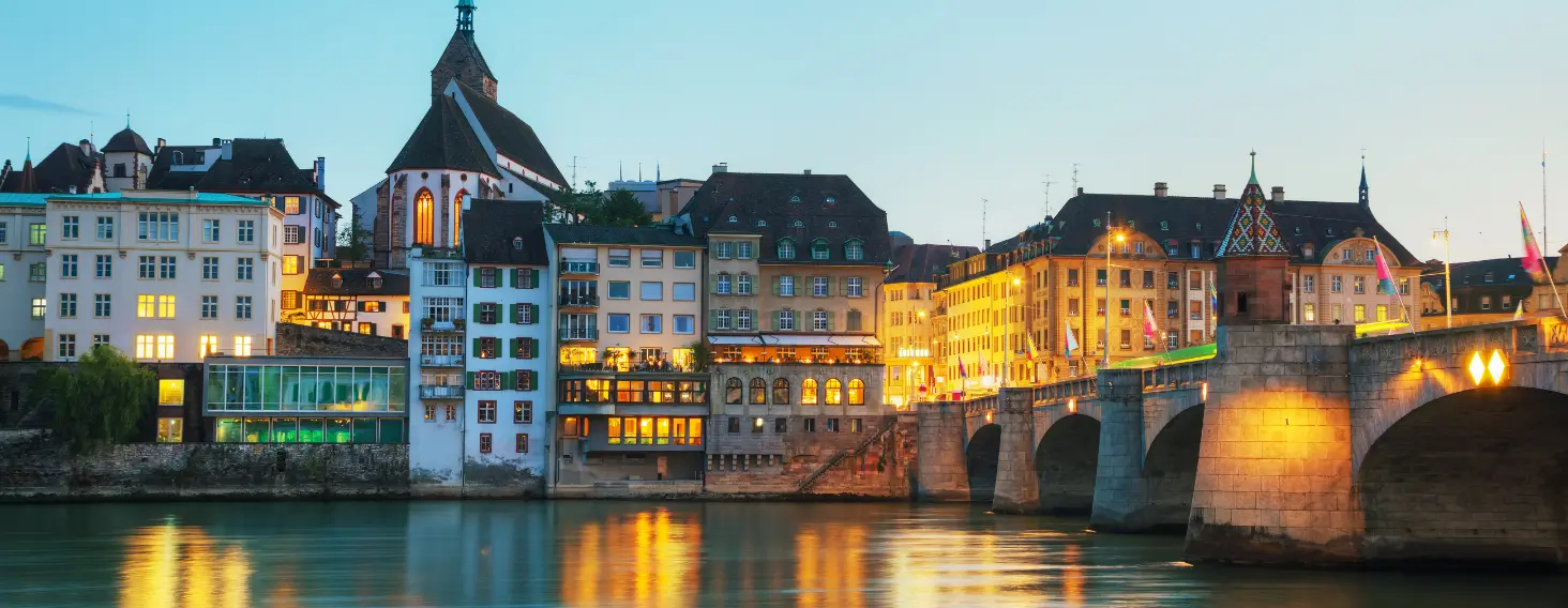 Colorful historic buildings along a calm river at dusk, with an illuminated stone-arch bridge reflecting in the water.