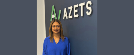 Portrait of a woman in a blue blouse standing in front of a dark blue wall with the company sign 'AZETS' above her left shoulder.
