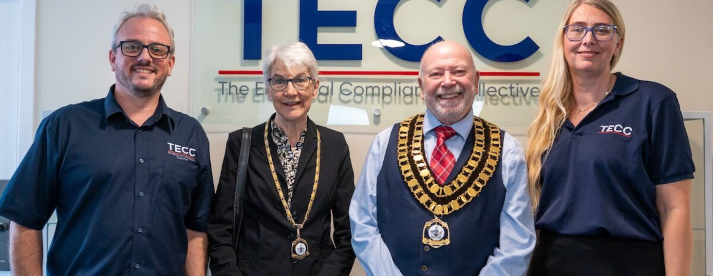 NEW - News item size 92 | Thames Valley Chamber of Commerce Four people pose in an office lobby in front of the TECC sign; two wear ceremonial chains and medallions, indicating official roles or honors.