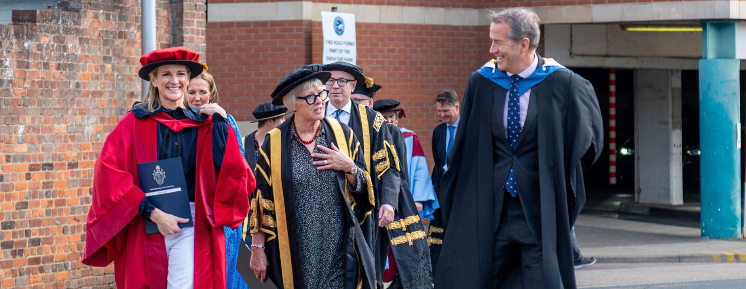 NEW - News item size 93 | Thames Valley Chamber of Commerce Group of academics in colorful ceremonial robes walking outdoors beside a brick wall, smiling and carrying folders for a procession event.