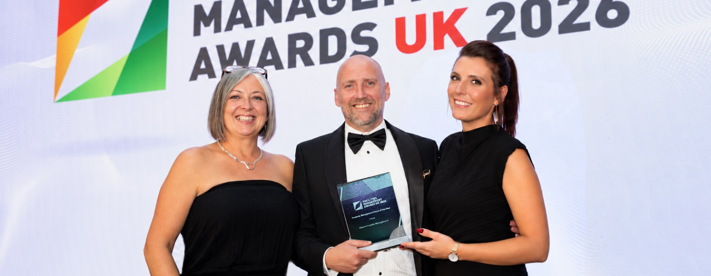 NEW - News item size 96 | Thames Valley Chamber of Commerce Three people pose on stage at an awards ceremony; a bald man in a tuxedo holds a glass award, flanked by two smiling women in black dresses.