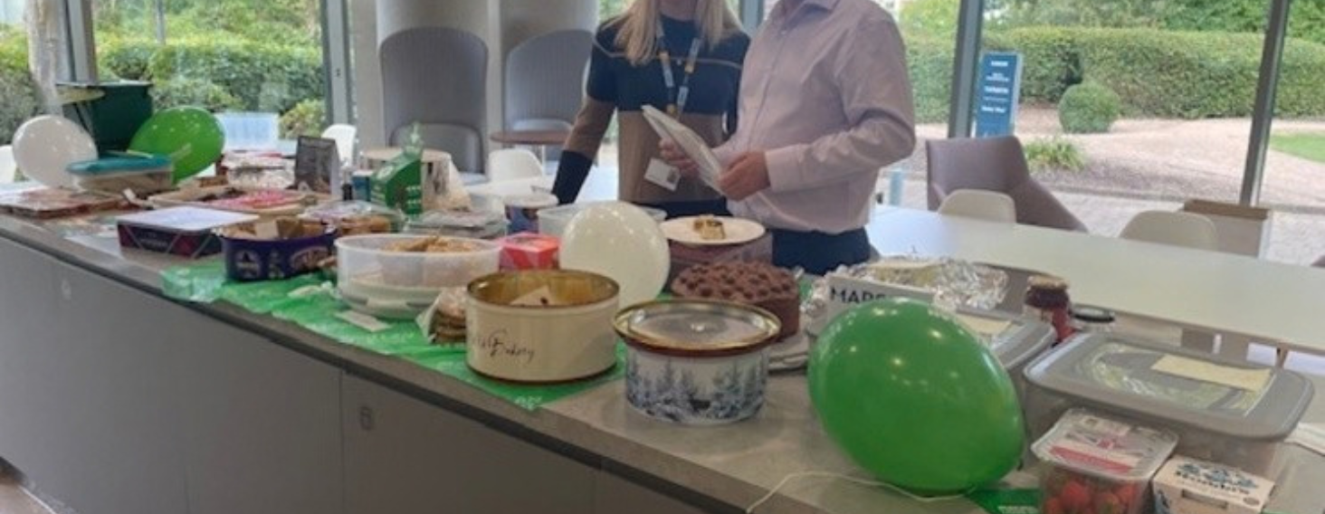 People standing behind a long table filled with desserts and snacks, with green and white balloons in a bright event space.