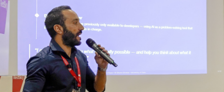 Man giving a talk into a handheld microphone at a conference, purple presentation slide in background.