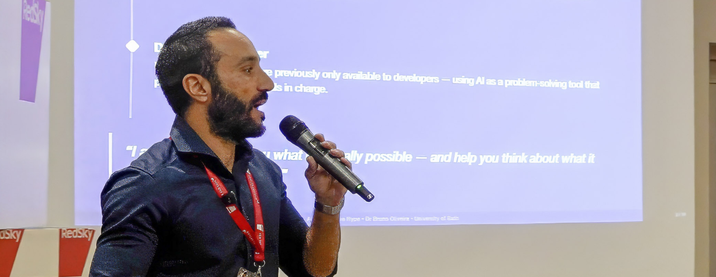 Man giving a talk into a handheld microphone at a conference, purple presentation slide in background.