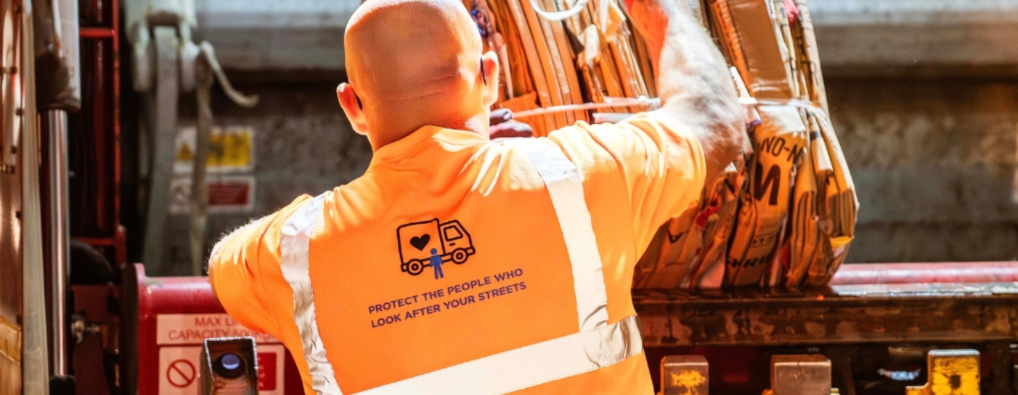Worker in orange safety vest loading bags into a truck; back shows a message about protecting street workers with a heart logo.