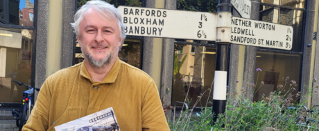 Smiling man with gray hair and beard, wearing a mustard shirt, standing beside a vintage town signpost.