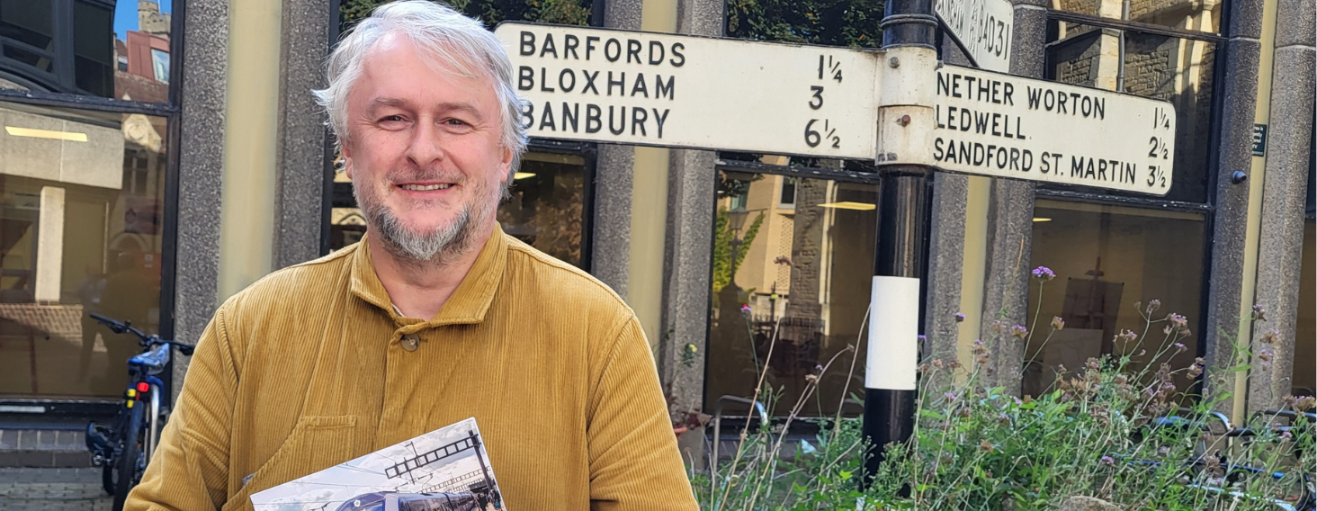 NEW - News item size - 2026-10-21T094122162 | Thames Valley Chamber of Commerce Smiling man with gray hair and beard, wearing a mustard shirt, standing beside a vintage town signpost.