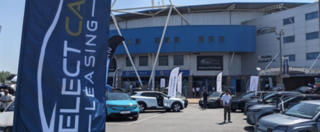 Outdoor car show in a parking lot with rows of cars on display and large blue banners.