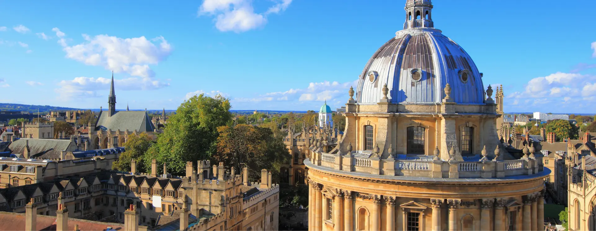 Aerial view of Oxford city with the Radcliffe Camera dome in the foreground and historic stone buildings under a blue sky.