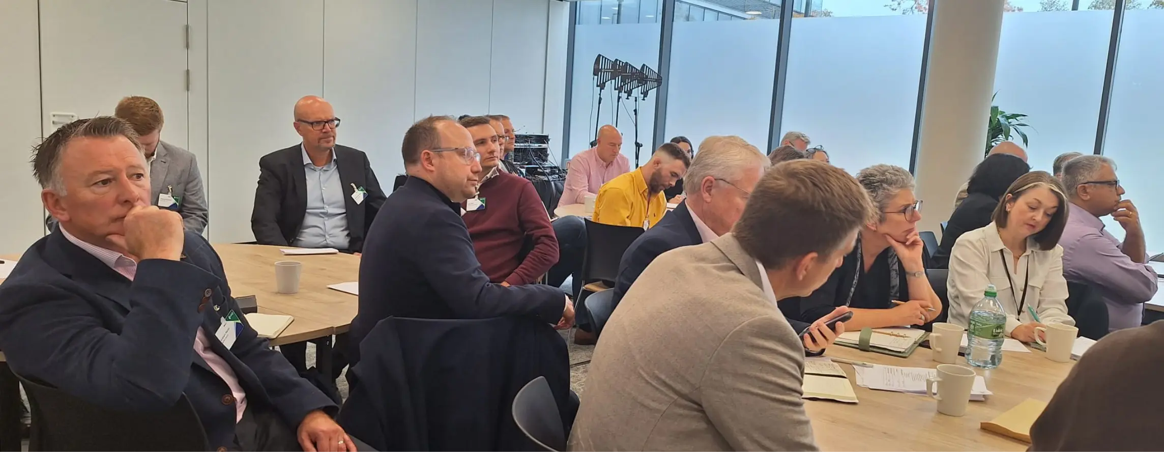 Group of professionals in a conference room seated around a long table, listening and taking notes.