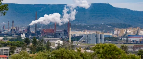 Industrial factory complex with tall smokestacks emitting white steam, set against green hills and trees in the foreground and background.
