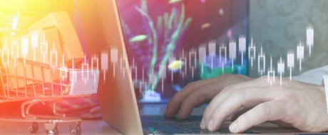 Hands typing on a laptop with a small shopping cart beside, overlaid by translucent stock charts in warm colors.
