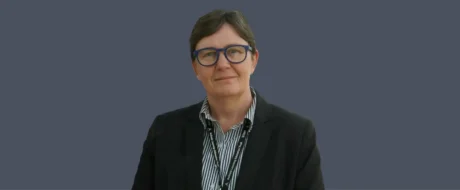 Professional staff portrait: woman with short gray hair, blue glasses, black blazer, striped shirt, and a staff lanyard against a solid blue-gray background.