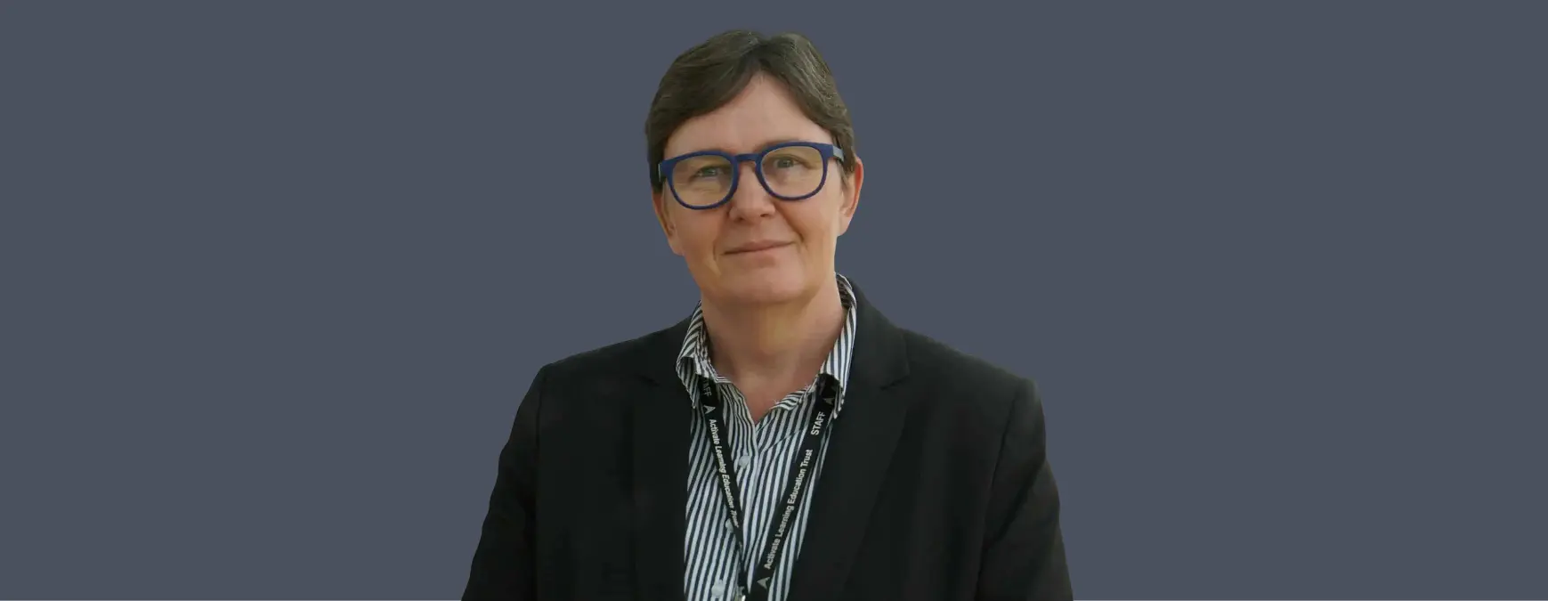 NEW - News item size 97 | Thames Valley Chamber of Commerce Professional staff portrait: woman with short gray hair, blue glasses, black blazer, striped shirt, and a staff lanyard against a solid blue-gray background.