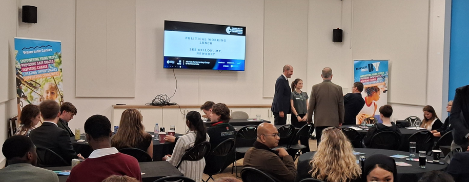 People networking at a luncheon event in a conference room; a large screen reads 'Political Working Lunch' on stage.