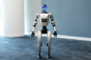 Robot Image.png | Thames Valley Chamber of Commerce Silver humanoid robot standing upright in a blue-carpeted showroom, facing the camera with a blue-lit face shield and a chest label reading'Extend Robotics'. merge to audience?