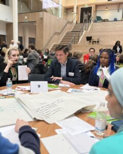 Velocity.jpg | Thames Valley Chamber of Commerce Diverse group seated around a round table in a modern atrium, discussing with papers and a large brainstorming sheet; a table number 9 visible.