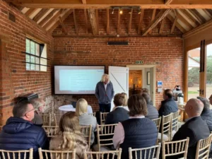 tv2.webp | Thames Valley Chamber of Commerce Speaker with long gray hair presents to a seated audience in a brick-walled room with a projector screen showing slides.